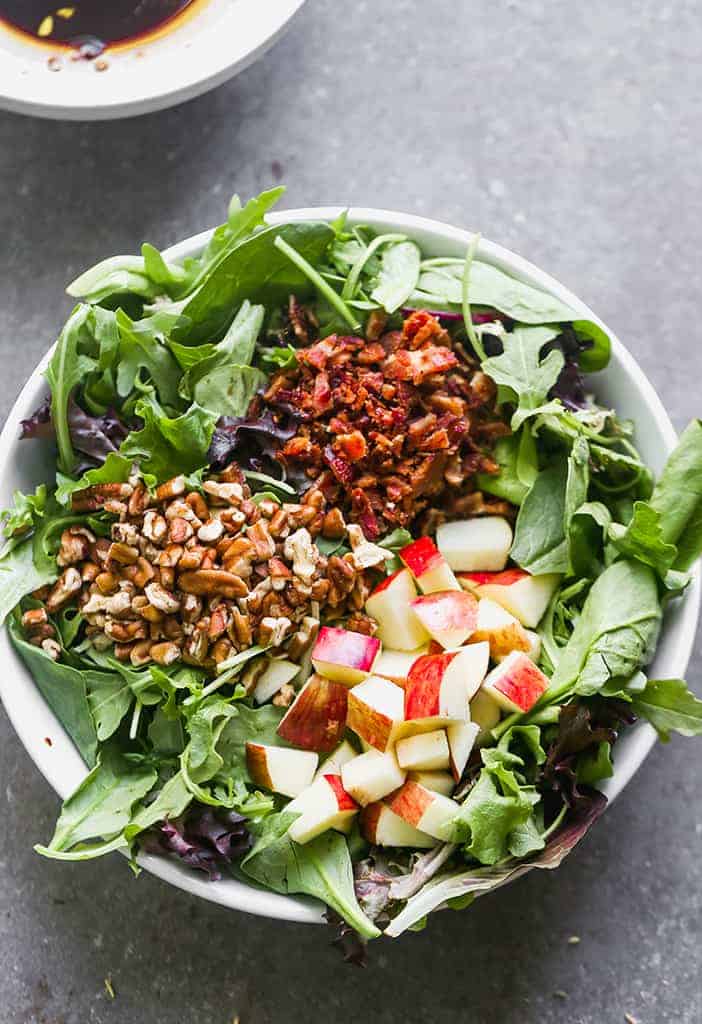 A green salad with three toppings laid in sections on top of the lettuce including chopped red apples, pecans and crumbled bacon.