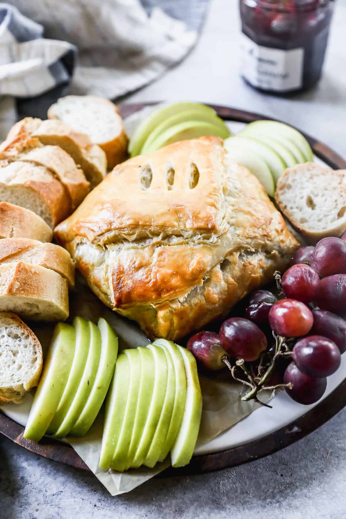 Baked Brie wrapped in a puff pastry on a platter surrounded by grapes, apples, and bread.