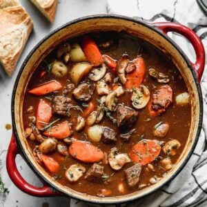 A large pot of beef stew with carrots, potatoes and mushrooms and sliced baguette on the side.