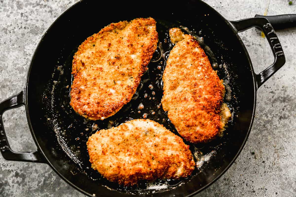 Three breaded chicken breasts being browned in a cast iron pan to make easy Chicken Parmesan.