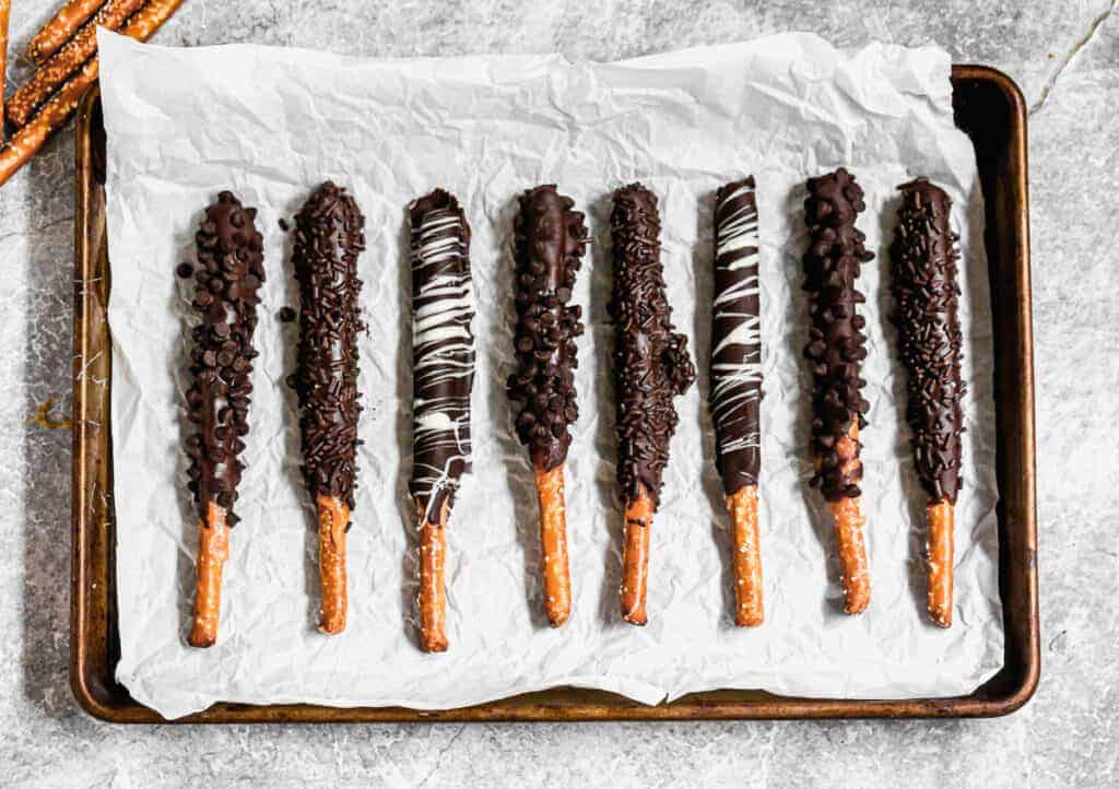 Chocolate dipped pretzel rods lined on a baking sheet, topped with mini chocolate chips, sprinkles, or a drizzle of white chocolate..