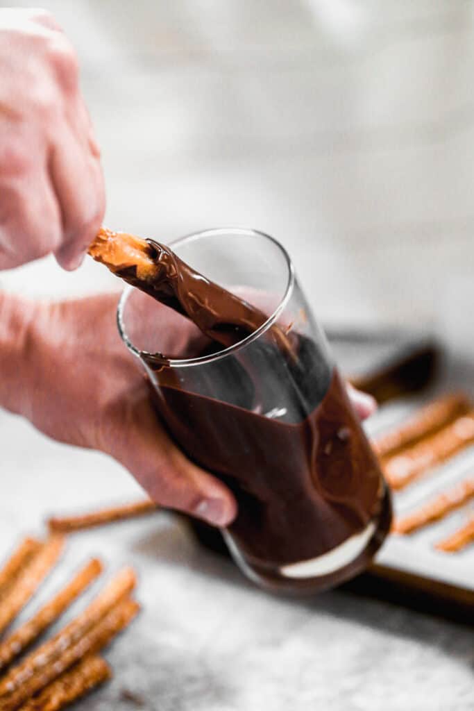 A hand dipping a pretzel rod in a cup of melted chocolate.