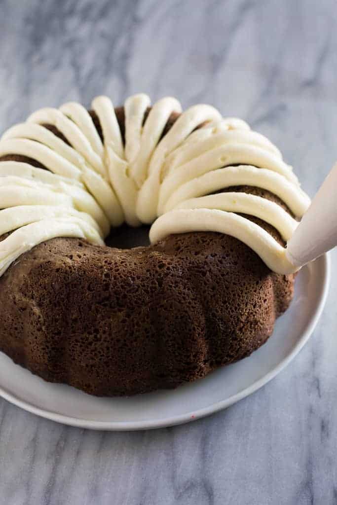 A chocolate bundt cake being frosted with cream cheese frosting using a pastry bag.