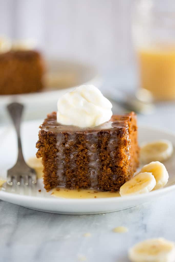 Gingerbread cake served on a white plate with a dollop of whipped cream on top, sliced bananas on the side and a vanilla cream sauce on top.