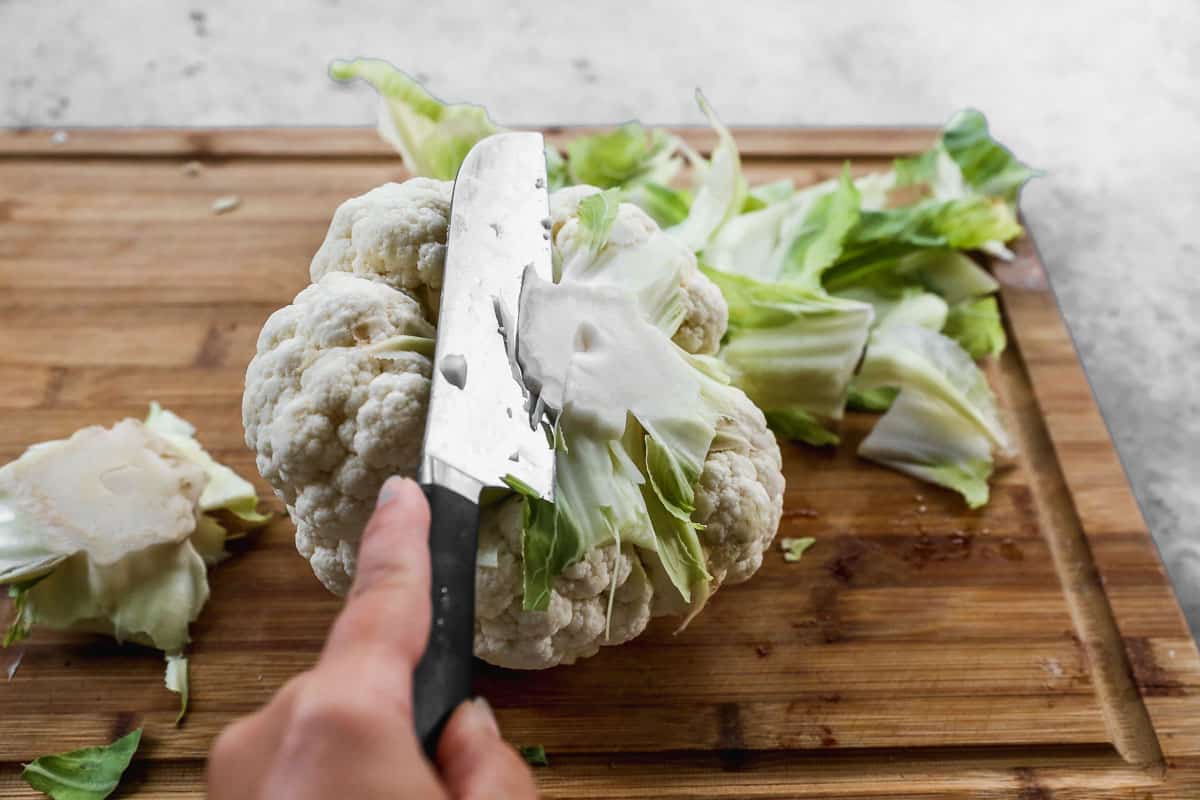 A large knife cutting the bottom off of a whole cauliflower.